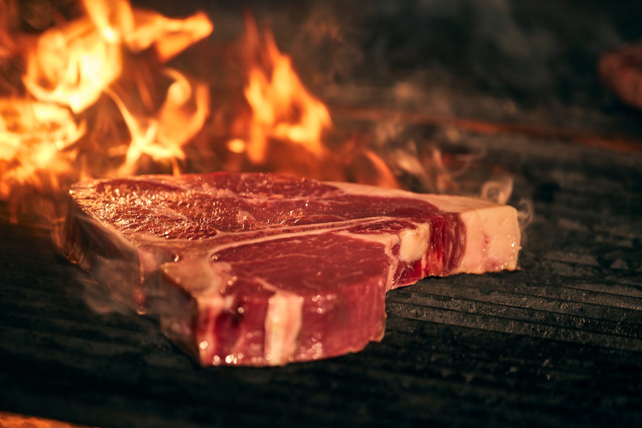 T-bone steak being grilled over open flames, showcasing sizzling outdoor barbecue cooking.